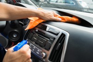 Person reinigt mit einem Lappen den Innenraum des Fahrzeugs und hält eine Sprühflasche mit passendem Reiniger in der Hand