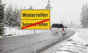 Fahrzeug auf verschneiter Straße, davor ein Ortseingangsschild auf dem Sommerreifen durchgestrichen ist und Winterreifen zu lesen sind