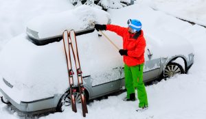 Zugeschneites Auto, dass vom Fahrer in Skikleidung freigeschaufelt wird. Am Auto sind seine Skier angelehnt.