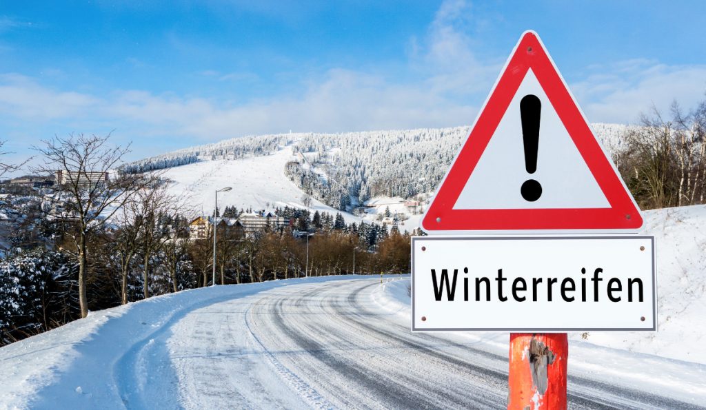Dreieckiges Warnschild mit einem Ausrufezeichen und darunter dem Hinweis auf Winterreifen auf verschneiten Straßen als Symbol für Winterreifen Pflicht