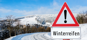 Dreieckiges Warnschild mit einem Ausrufezeichen und darunter dem Hinweis auf Winterreifen auf verschneiten Straßen als Symbol für Winterreifen Pflicht